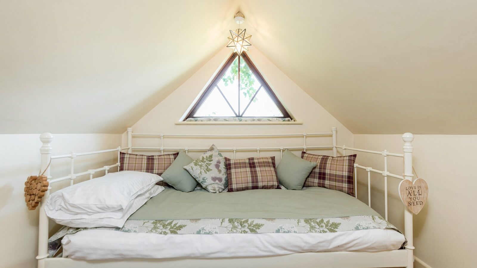 Cozy attic bedroom with a white metal daybed, green and plaid pillows, and a triangular window adorned with a star-shaped light above—bringing a touch of Lavender Hill Treehouses charm to the space.