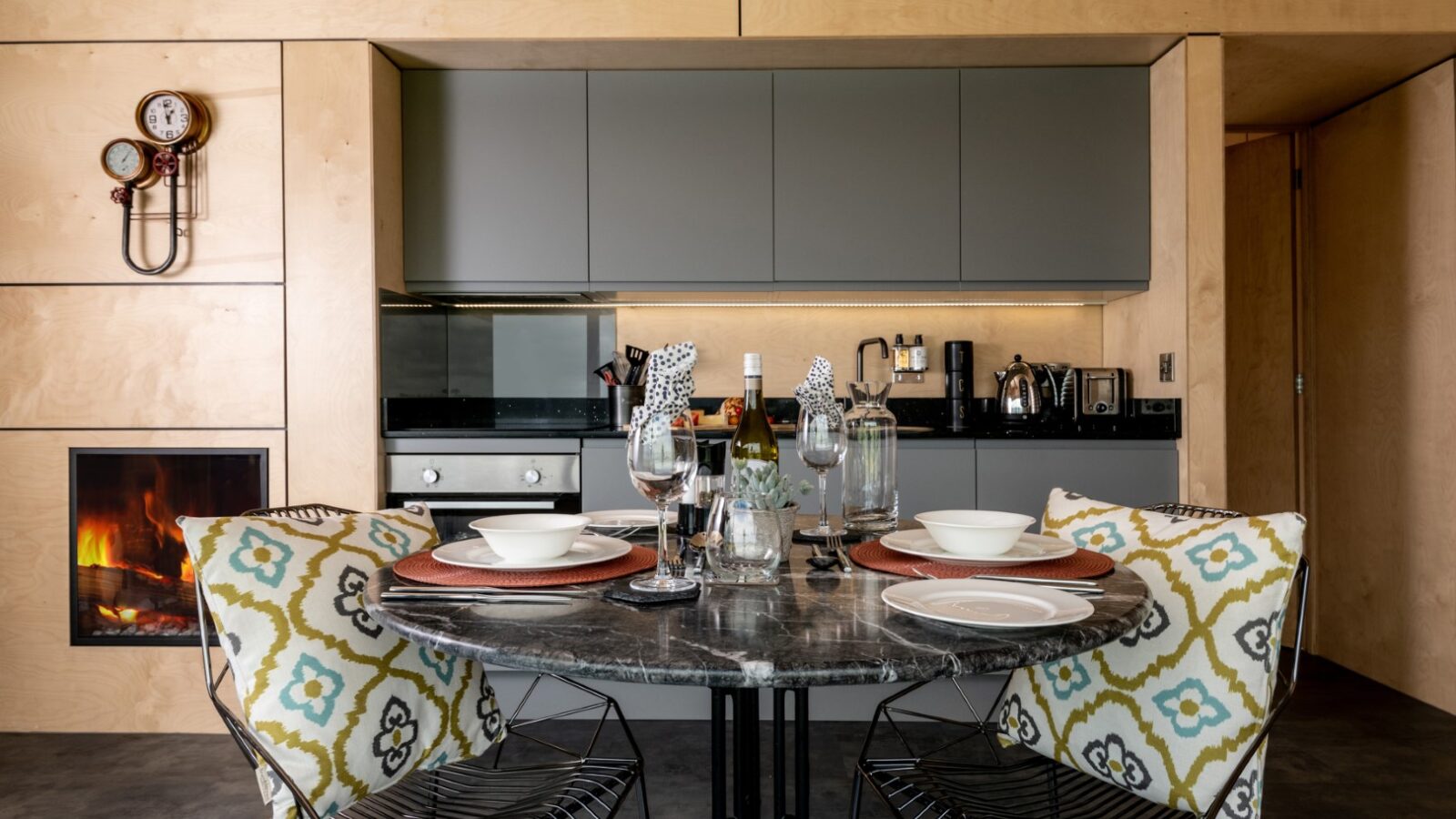 Modern kitchen at Bettws Court Retreats, featuring a round table set for two with wine glasses and patterned cushions on chairs. Fireplace and cooking area are also visible, offering a perfect blend of comfort and style.