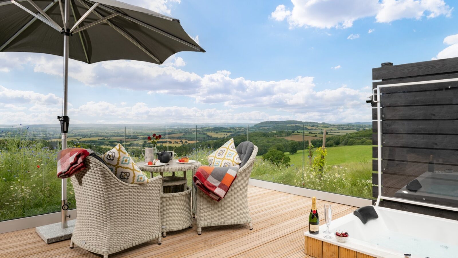 The outdoor patio at Bettws Court Retreats features wicker chairs, a table with refreshments, an umbrella, and a hot tub overlooking a scenic landscape.