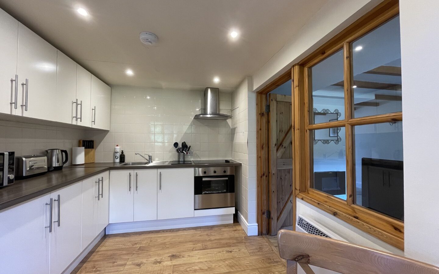 Modern kitchen with white cabinets, stainless steel appliances, and wooden flooring, reminiscent of the charming style found in Trallwm Forest Cottages. A wooden-framed glass window on the side completes this delightful space.