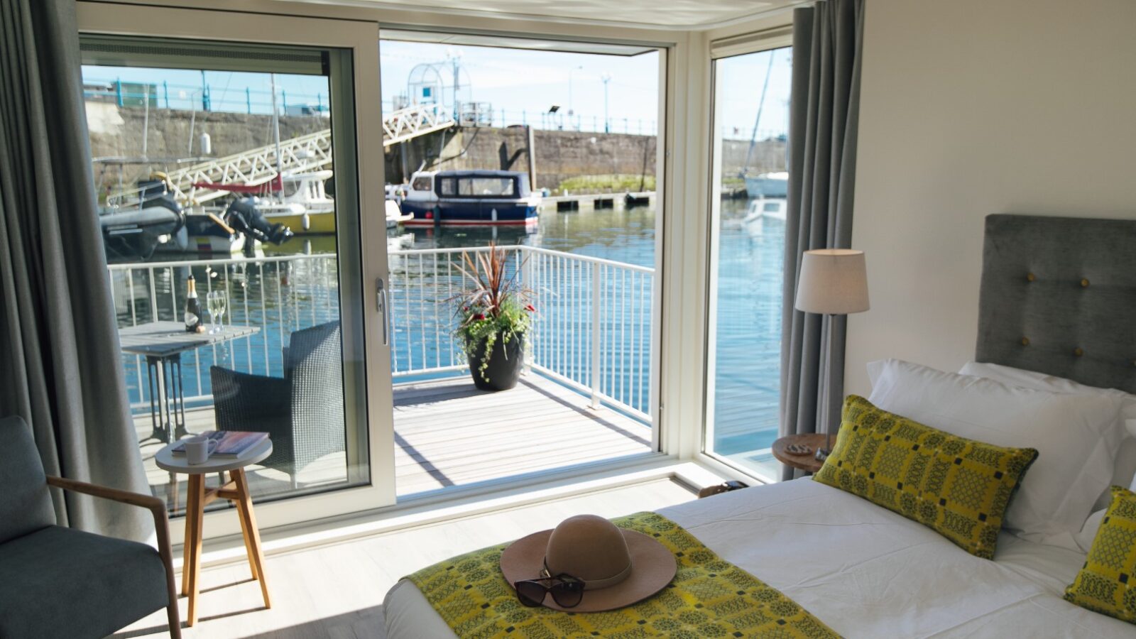 Bright bedroom with a double bed facing a glass door leading to a balcony overlooking Ty Milford Floatels, offering views of the marina with boats and a bridge.