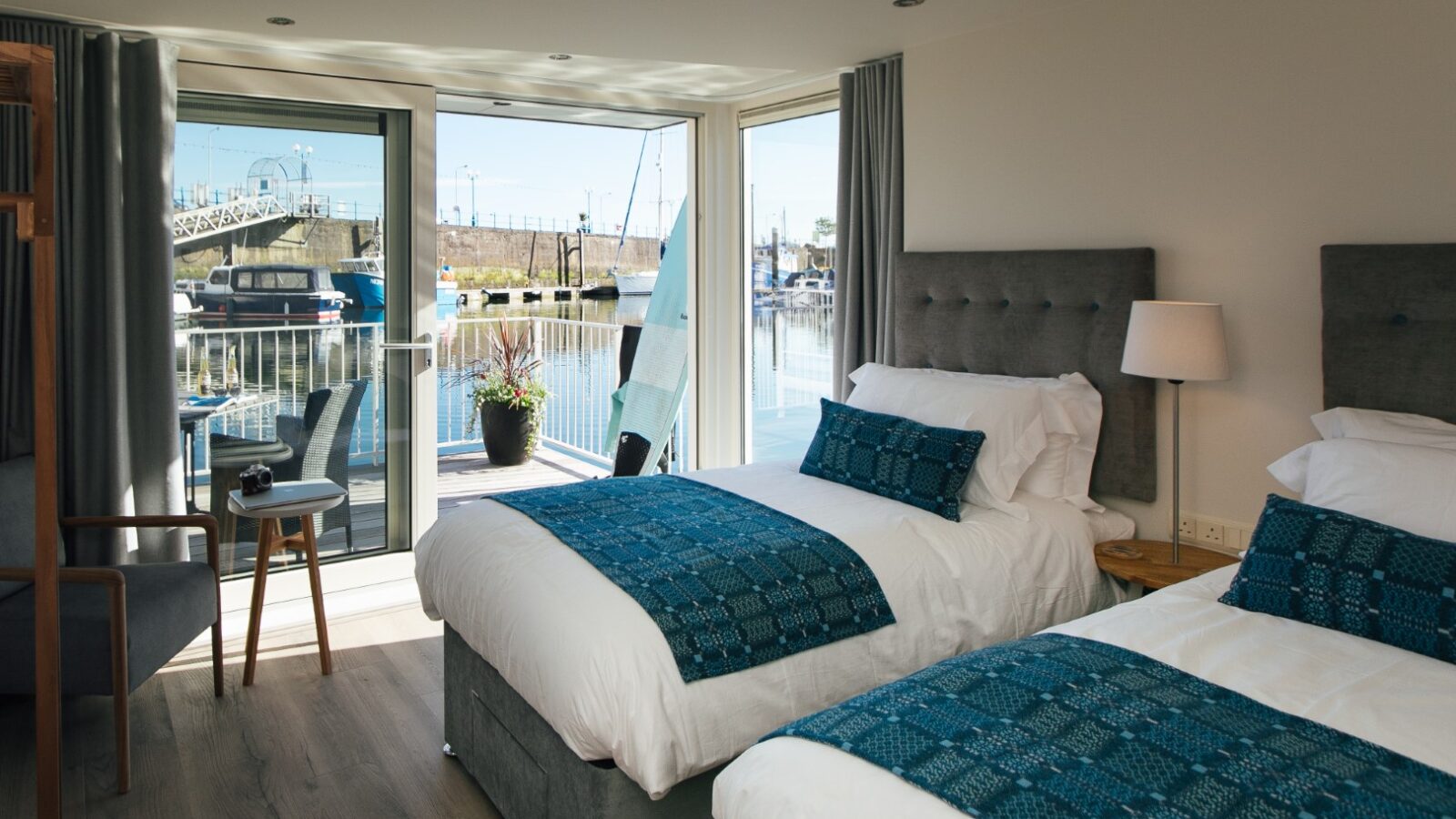 Experience a modern floatel room with two beds, blue decor, and a chair by the window. Enjoy breathtaking views of the harbor outside, reminiscent of the elegant style typified by Ty Milford's designs.