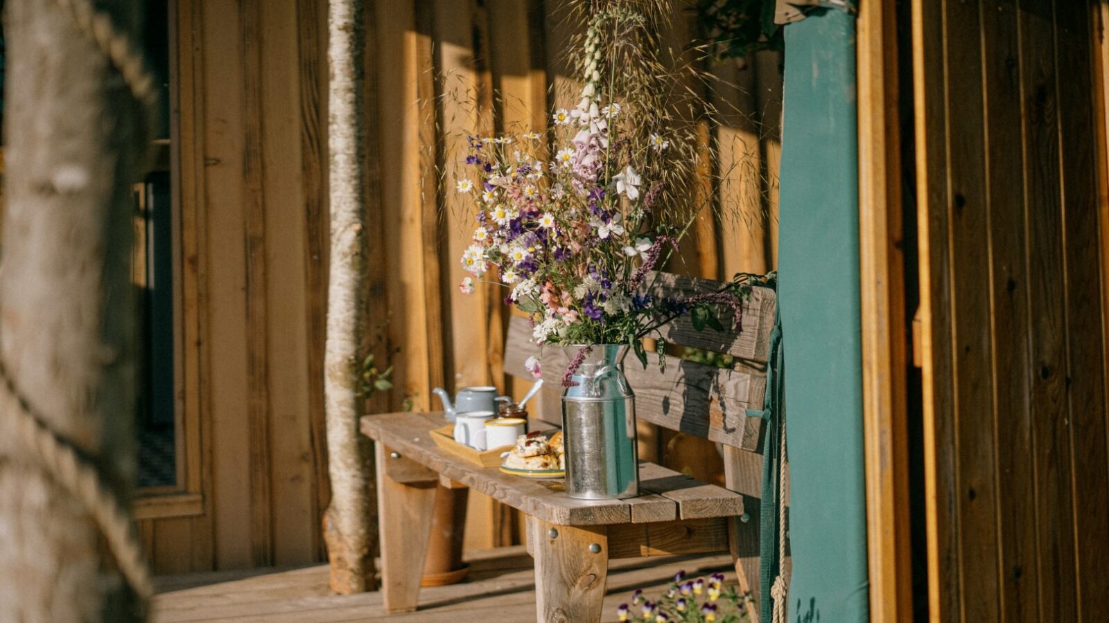 A wooden bench sits on a deck outside a rustic cabin at Southleigh Farm, adorned with a large, colorful bouquet of wildflowers in a tin vase. Nearby, cups, a teapot, and plates with food suggest a quiet, scenic spot for relaxation and tea amidst the charm of glamping in the Meadow Yurt.