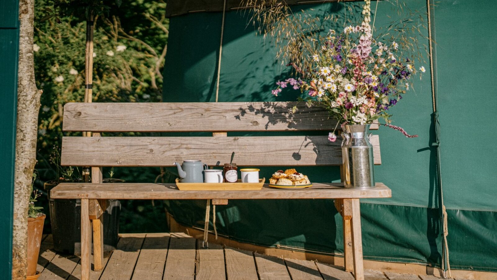 A rustic wooden bench sits on a wooden deck at Southleigh Farm. On the bench, there's a tray with a kettle, cups, and a jar of preserves, along with pastries. A tall bouquet of colorful wildflowers in a metal vase is placed beside the tray. The background features a charming Meadow Yurt.