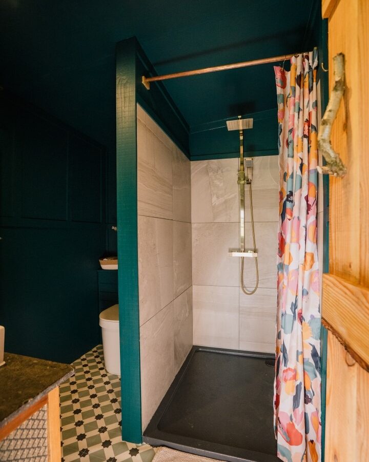 A bathroom with dark teal walls at Southleigh Farm features a shower area with beige tiles, a modern overhead shower fixture, and a colorful floral shower curtain. The floor has patterned black and white tile, and a wooden door is partially visible on the right.