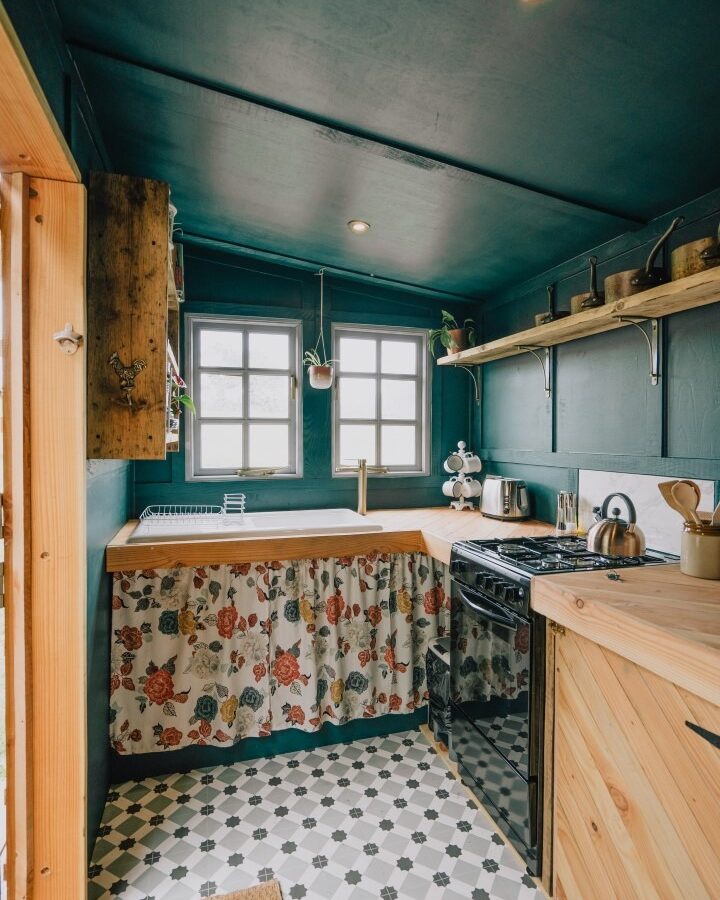 A cozy kitchen at Southleigh Farm features wooden countertops, a stove, and an oven. Floral-patterned curtains cover the storage space beneath the sink. Shelves holding kitchen items are mounted above. Two windows let in natural light, and the floor is decorated with black and white tiles.