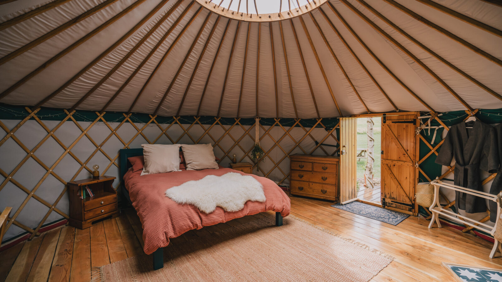 Inside the Meadow Yurt at Southleigh Farm, a cozy haven awaits. A round skylight crowns the space, illuminating a rust-colored duvet atop the neatly made bed. A soft rug adorns the wooden floors, while rustic furnishings enhance the charm. Light streams through an open door, highlighting tranquility.