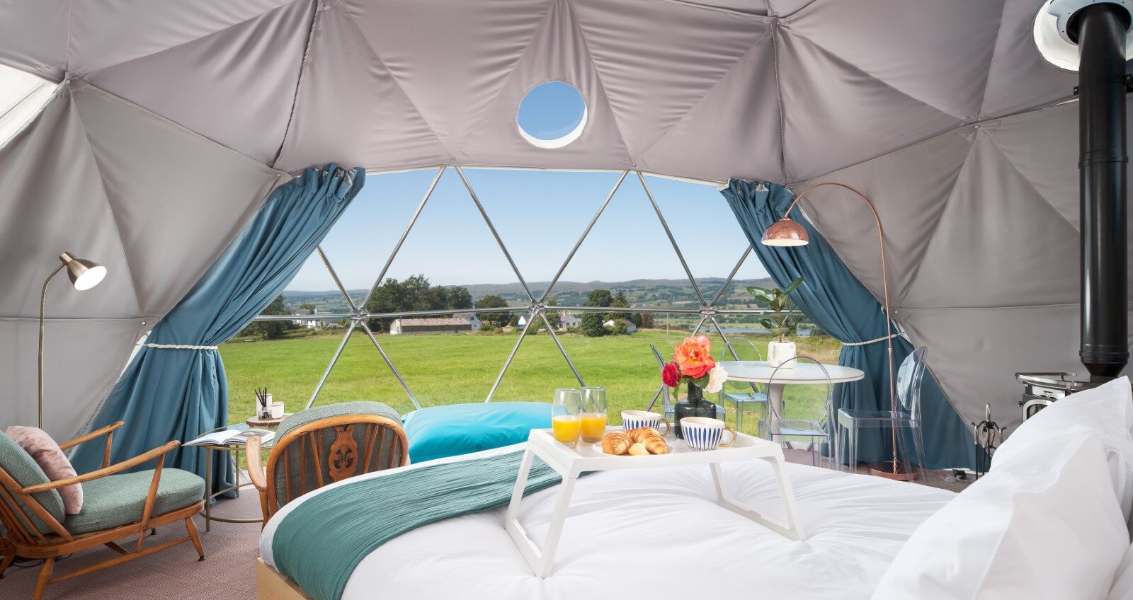 The interior of the Mid Auchengowan Dome glamping tent features a cozy bed, a breakfast tray, and chairs, all perfectly framed by the large windows offering a stunning view of lush green fields.