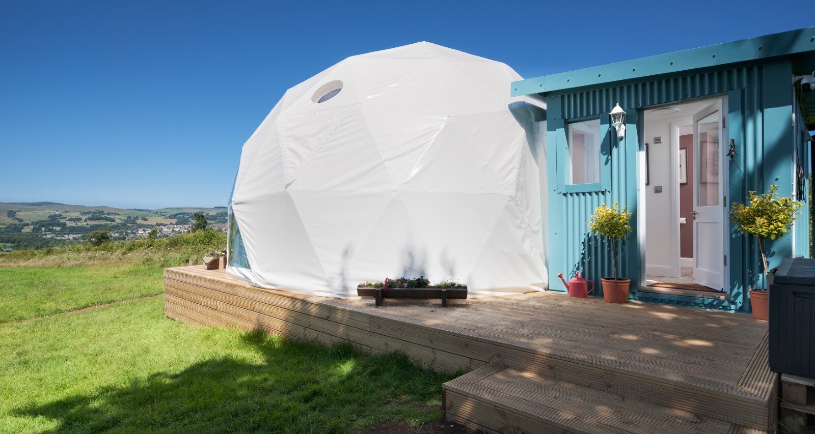 The Mid Auchengowan Dome, a white geodesic structure, and a small blue cabin are perched on a wooden deck in a grassy field under the expansive, clear blue sky.