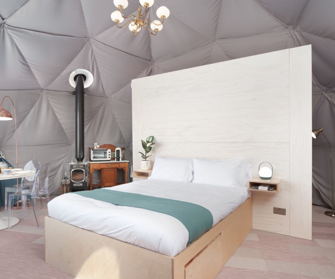 The Mid Auchengowan Dome features a modern interior with a cozy bed draped in white bedding, complemented by a wooden panel headboard. A vintage radio sits quaintly by the stove, while the chandelier lighting adds an elegant touch to this inviting dome sanctuary.