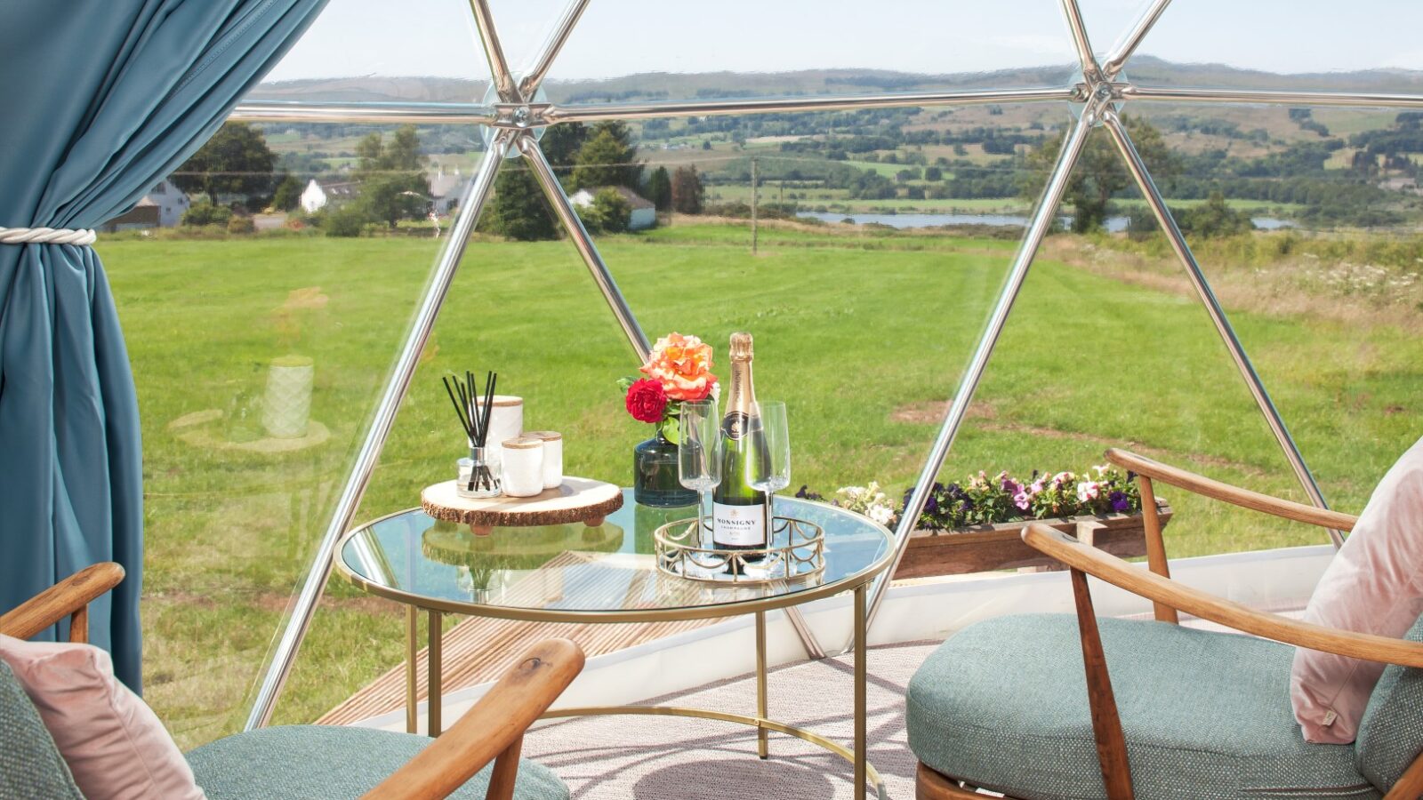 The Auchengowan Dome's interior features a chic table adorned with champagne, glasses, and fresh flowers, offering a luxurious glamping experience. Through the expansive window, guests can enjoy breathtaking views of the lush green landscape.