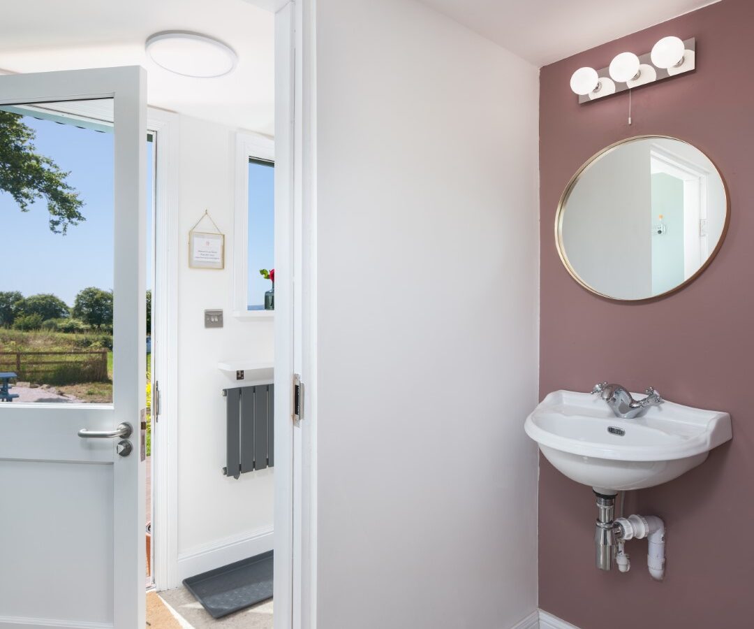 A small bathroom with a wall-mounted sink and round mirror reflects the Mid Auchengowan Dome's charm. Through the open door, a sunny outdoor scene bathes the space in natural light.