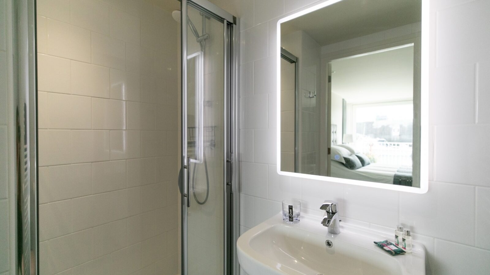 Modern bathroom with a glass-enclosed shower, white sink, and illuminated mirror by Ty Milford. A glimpse of the bedroom is visible in the mirror reflection.