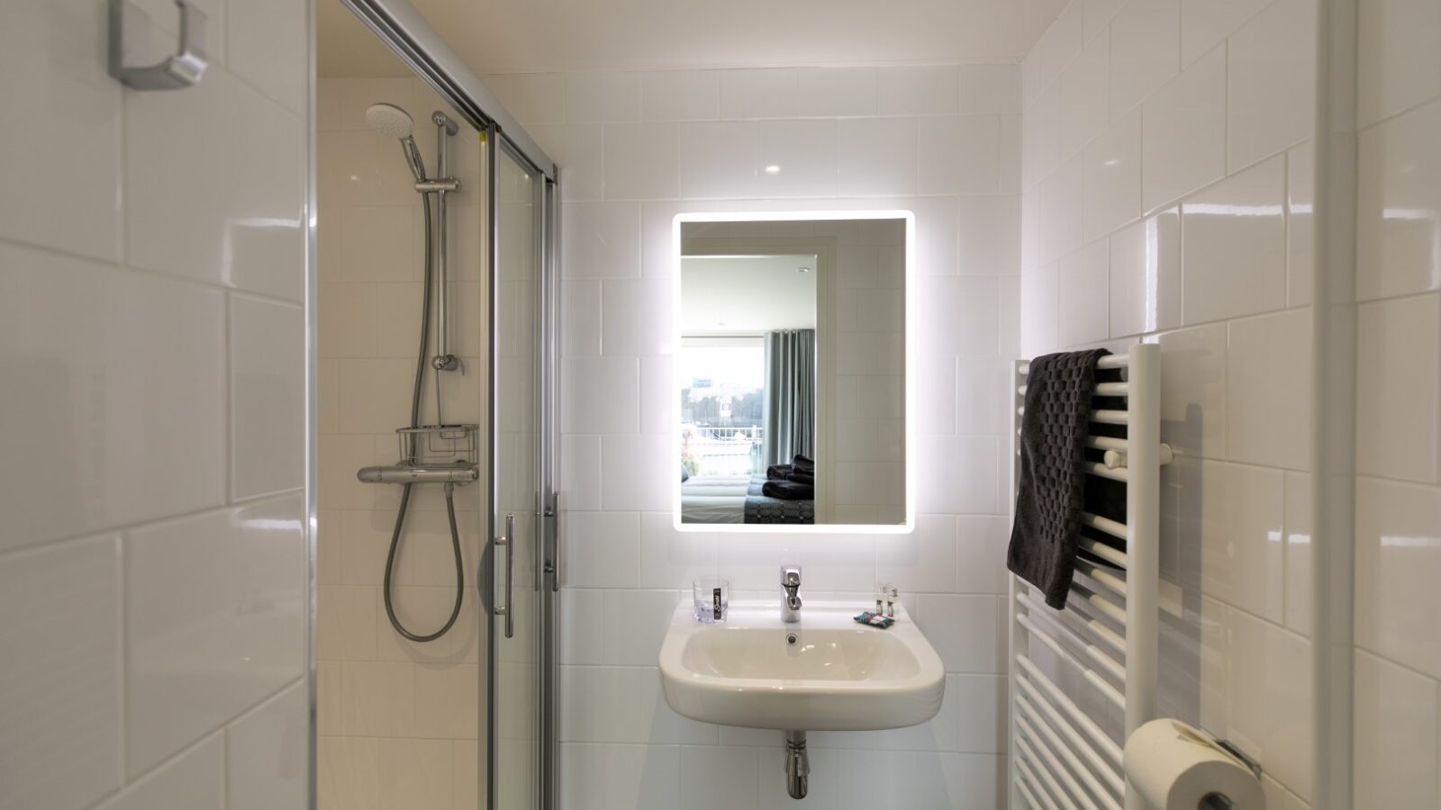 Experience a modern bathroom at Ty Milford Floatels, featuring a sleek glass shower, illuminated mirror, pristine white sink, radiator, and towel rack set against elegant tiled walls. Enjoy the perfect blend of comfort and style at this unique waterfront accommodation.