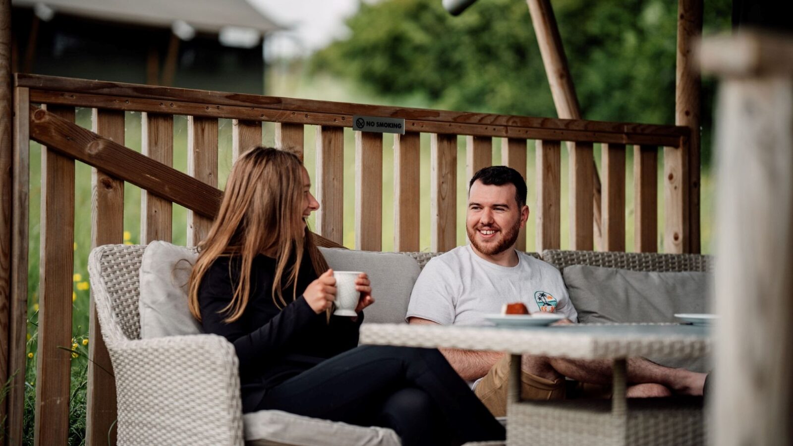 A man and woman sit on a wicker sofa outdoors, enjoying coffee amidst nature. The woman wears a black outfit and the man sports a white T-shirt. They are smiling and engaged in conversation, nestled in this serene setting with a wooden fence enclosing the area.