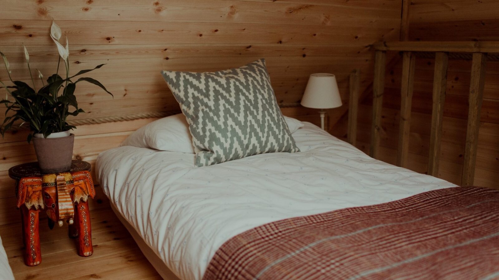 A cozy bedroom in the Netherby Treehouse with wooden walls and flooring features a single bed. The bed has a white duvet, a gray-patterned pillow, and a red checkered blanket partially covering it. Beside the bed is an ornate stool holding a potted plant, and a lamp on the other side.