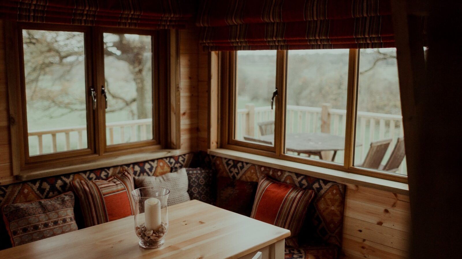 A cozy wooden cabin interior reminiscent of the Netherby Treehouse, featuring a dining table with an ornate candle holder, surrounded by patterned cushions on bench seats. Large windows offer a view of an outdoor deck with chairs and a table, overlooking a serene landscape of trees and fields.