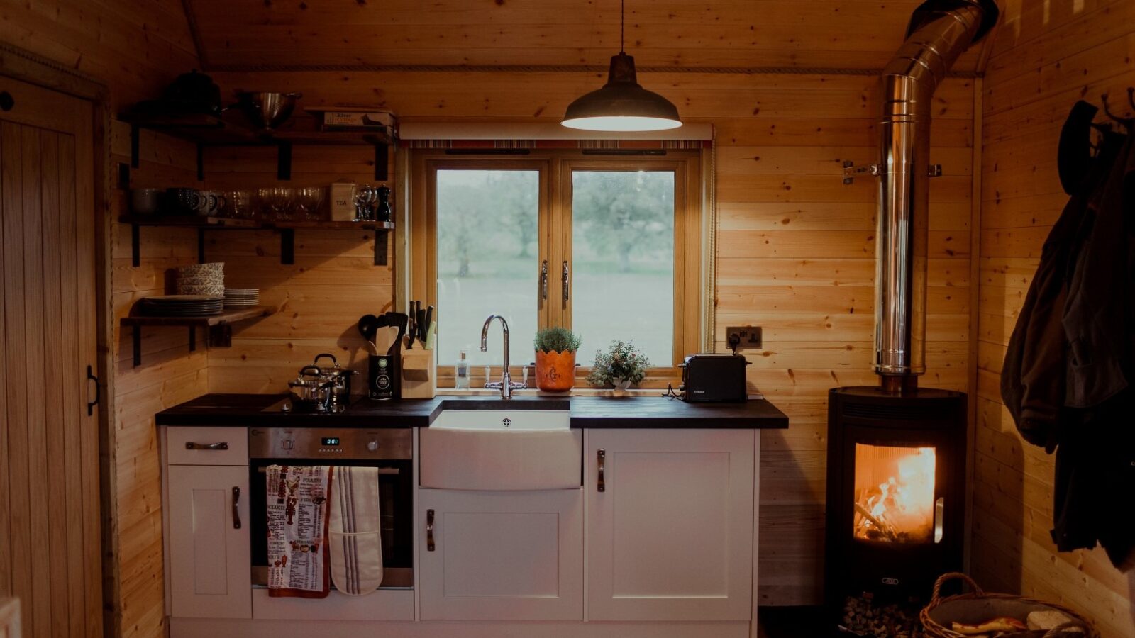 A cozy wooden kitchen at the Netherby Treehouse with a large window above a white farmhouse sink. To the right, a black wood-burning stove with a visible fire adds warmth. Shelves with utensils and appliances, including a coffee maker and toaster, are arranged neatly on the left wall of this unique lodging.