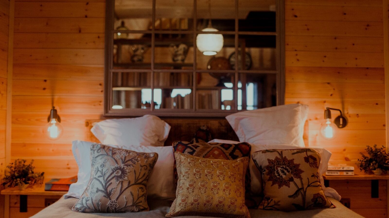 A cozy bedroom in the Netherby Treehouse with wooden walls features a neatly made bed adorned with patterned pillows. Two bedside tables hold small decorative items, and warm lighting from two wall-mounted lamps creates a welcoming ambiance. A large framed mirror hangs above the bed.