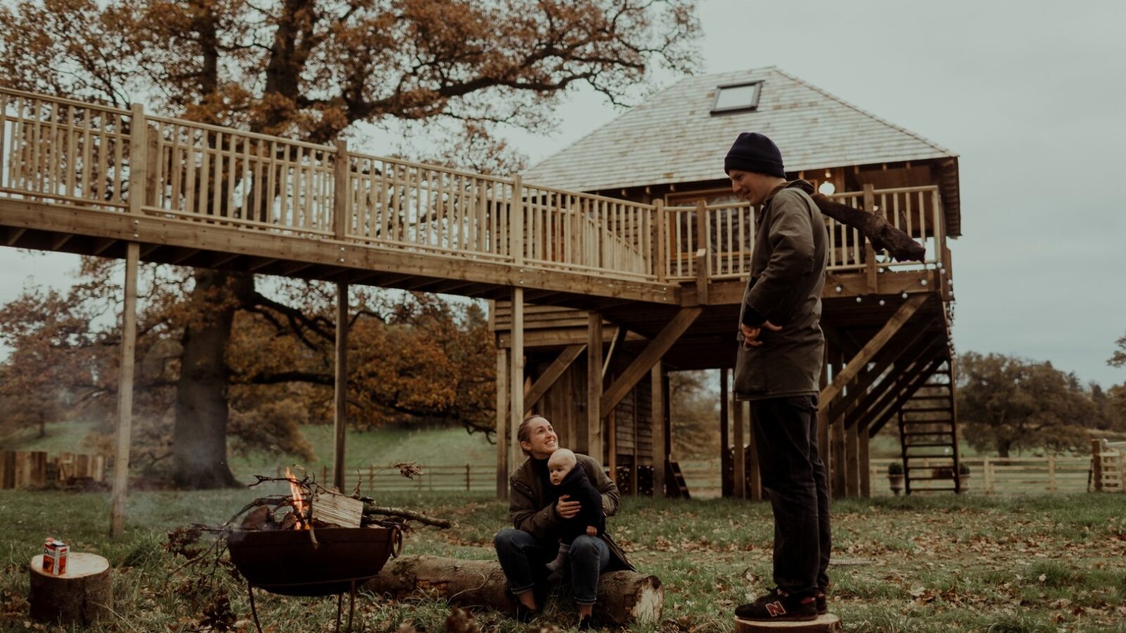 A man and a child are outside near a fire pit, surrounded by a grassy area with autumn leaves. The child sits on a log, wrapped in a blanket, while the man stands nearby. In the background, the charming Netherby Treehouse rental beckons with railings and a ladder, perfect for an unforgettable vacation rental experience.