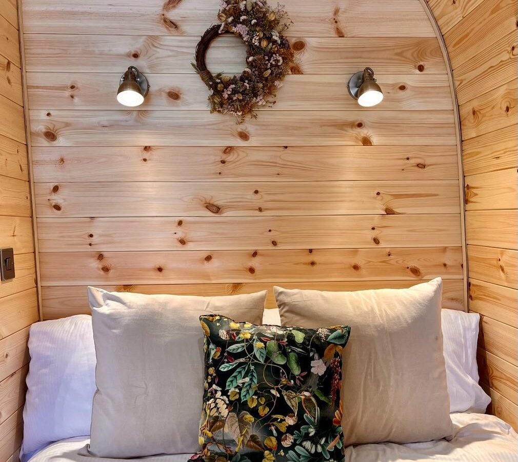 Cozy bedroom with wood-paneled walls, featuring a bed adorned with white pillows and a decorative pillow with a floral design. Above the bed, two wall-mounted lamps and a dried flower wreath add a touch of rustic charm—perfect for your New Farm glamping experience.