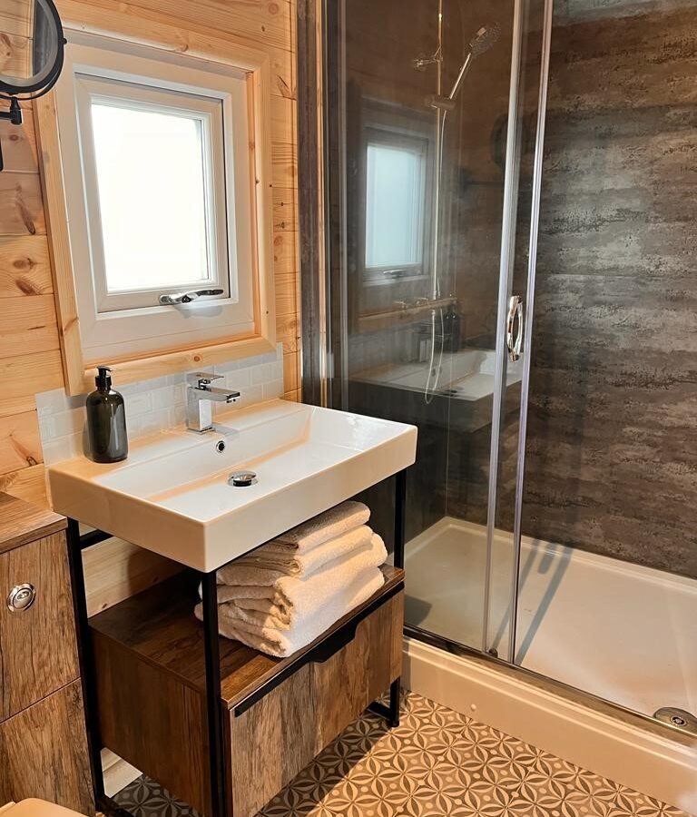 A modern bathroom with a wooden theme featuring a glass-enclosed shower, white sink with a black faucet, and a countertop with neatly folded white towels underneath. A small window above the sink allows natural light in, and patterned floor tiles add visual interest, echoing the charm of farm glamping.