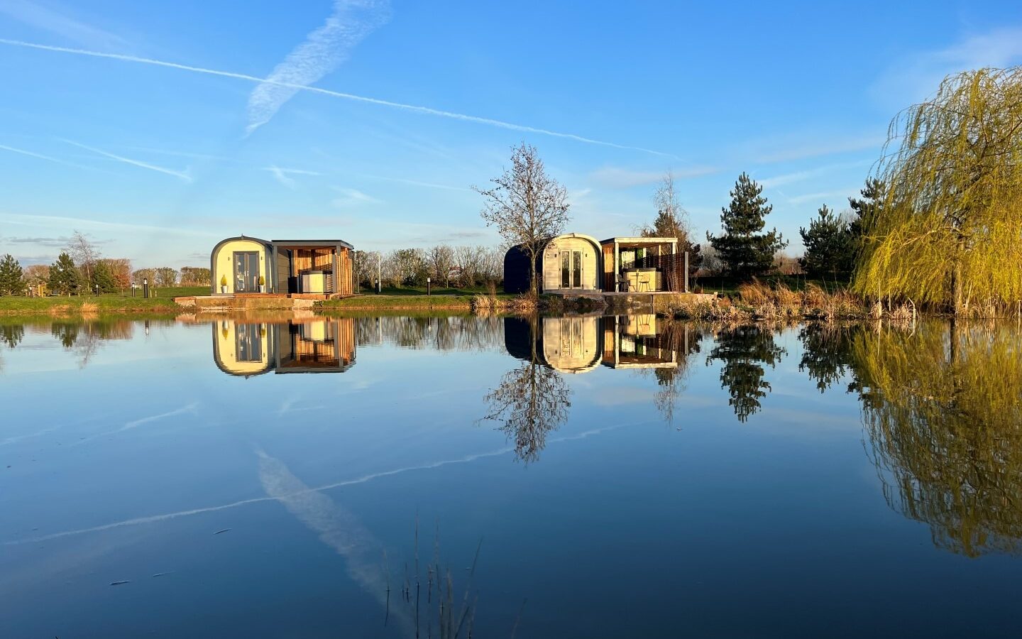 A tranquil lakeside scene features a cluster of new, modern cottages ideal for glamping, with trees reflected in calm water. The sky is clear with visible contrails, creating a serene and picturesque environment reminiscent of a peaceful farm setting.