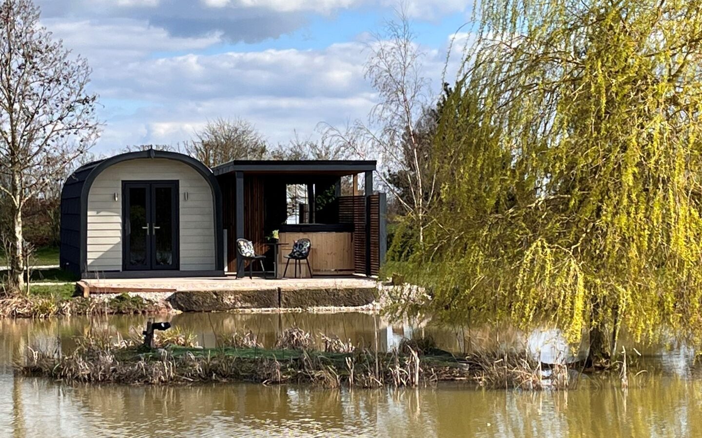 A modern, small cottage with arched roofs sits on the edge of a serene pond, surrounded by trees and greenery. This new glamping retreat features a front patio with chairs and a table. The sky is partly cloudy, casting a peaceful ambiance over the scene.