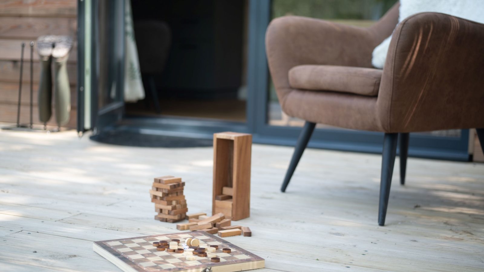 The outdoor wooden deck of Pond View Lodges features a brown armchair and open glass door. A chessboard and scattered wooden blocks on the floor add to the relaxed, casual atmosphere, perfect for unwinding with a serene pond view.