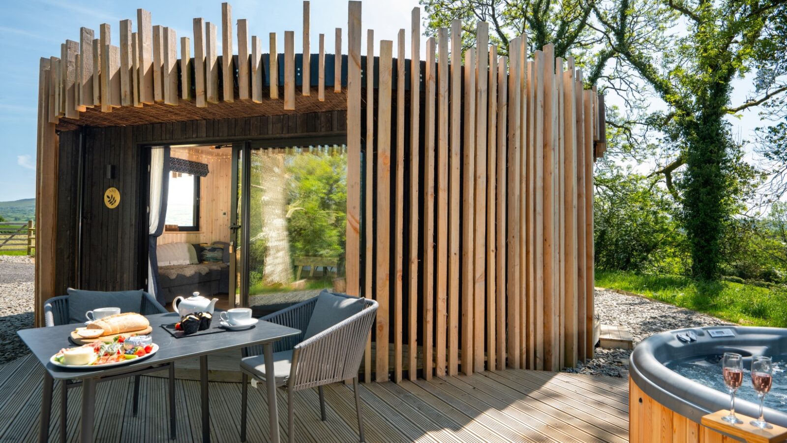 A modern wooden cabin with vertical slats, a glass front, and a deck overlooks the stunning Pond View. On the deck, a table is set with breakfast items and two chairs. A hot tub invites relaxation amidst the lush greenery and towering trees under a clear blue sky.