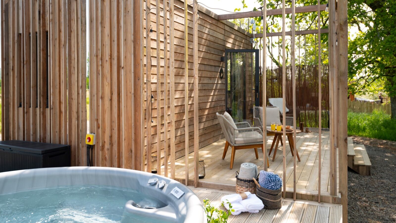 A wooden cabin lodge with vertical slats features a cozy outdoor seating area with two chairs and a small table. In the foreground, a hot tub is bubbling, offering a serene pond view. A basket with towels and a cozy blanket sits nearby, surrounded by greenery and sunlight.