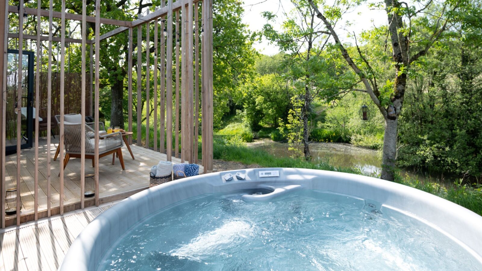 A bubbling hot tub sits on a wooden deck of the lodge, overlooking a lush, green forest. Two chairs and a table are placed on the deck, partially enclosed by wooden slats. Sunlight filters through the trees, highlighting the serene pond view in the background.