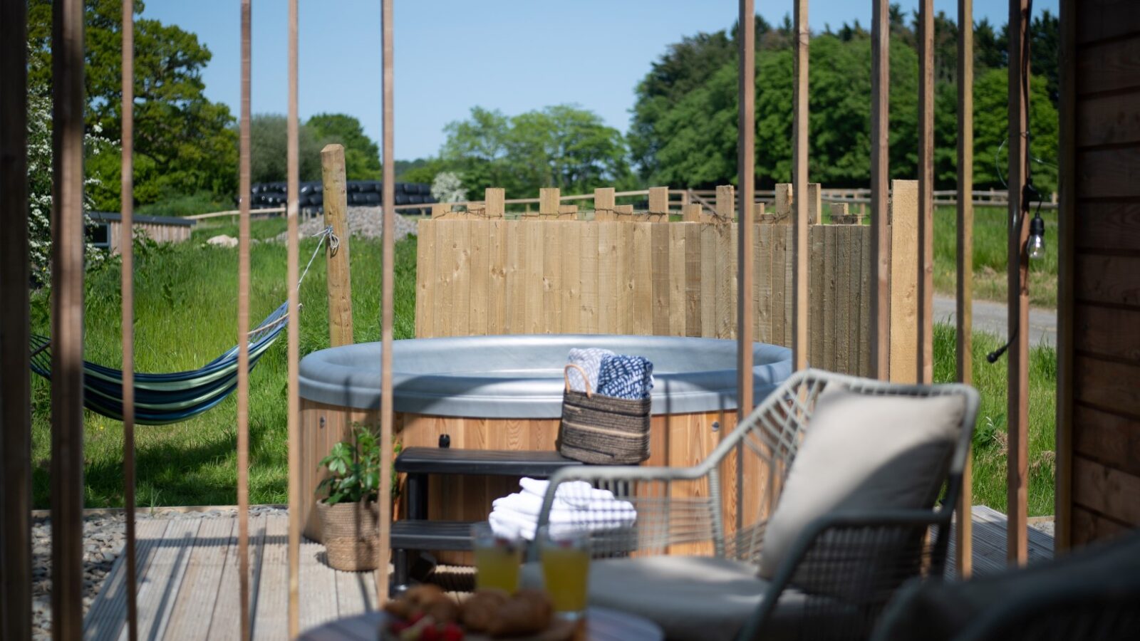 A cozy outdoor space at Pond View Lodges features a hot tub on a wooden deck, surrounded by greenery. A hammock is strung between posts, and a small table holds snacks and drinks. A chair with cushions offers seating in the foreground, all perfectly complementing the tranquil pond views.