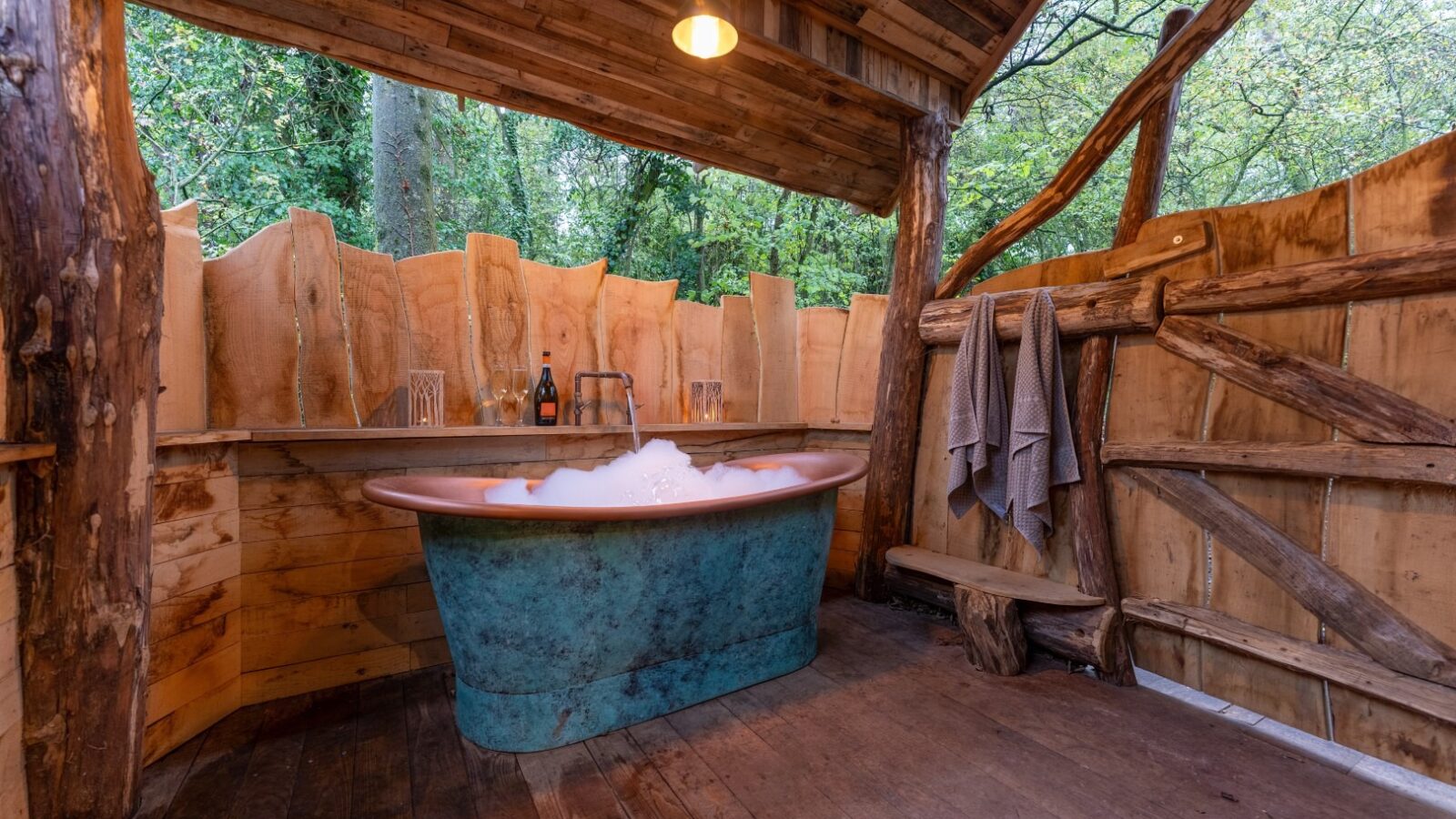 In the heart of One Acre Wood, discover an outdoor rustic bathroom featuring a teal bathtub filled with bubbles and surrounded by wood paneling. Towels hang from a log rack, while wine and glass rest on a wooden shelf. Trees frame the scene, illuminated by cozy lighting above.