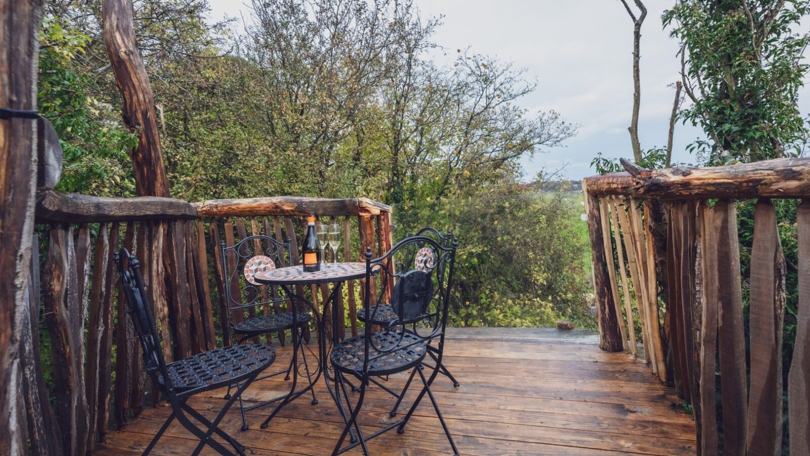 A small wooden balcony in One Acre Wood features a black wrought-iron table and chairs. On the table, there's a bottle of wine and two wine glasses, suggesting a cozy and serene setting for enjoying a drink while overlooking the forested landscape.