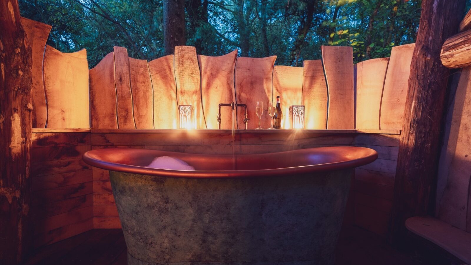 A rustic outdoor bath setup in One Acre Wood features a large metal tub filled with water and bubbles. Candles and lanterns are arranged around the tub, providing warm light. Wooden planks form a partial enclosure, and trees are visible in the background, creating a serene atmosphere.