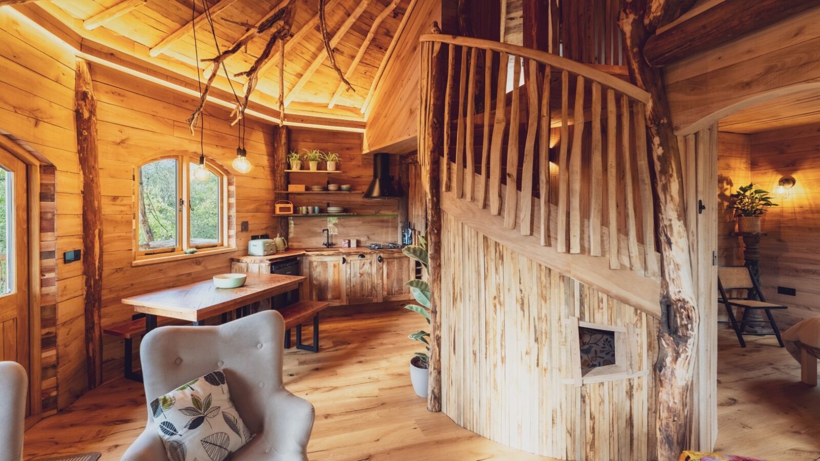 A cozy wooden cabin interior in the heart of One Acre Wood features a winding staircase made from natural wood. The space includes a sitting area with a cushioned chair, a plant, a kitchen with modern appliances, and large windows allowing ample natural light. Various plants decorate the space.