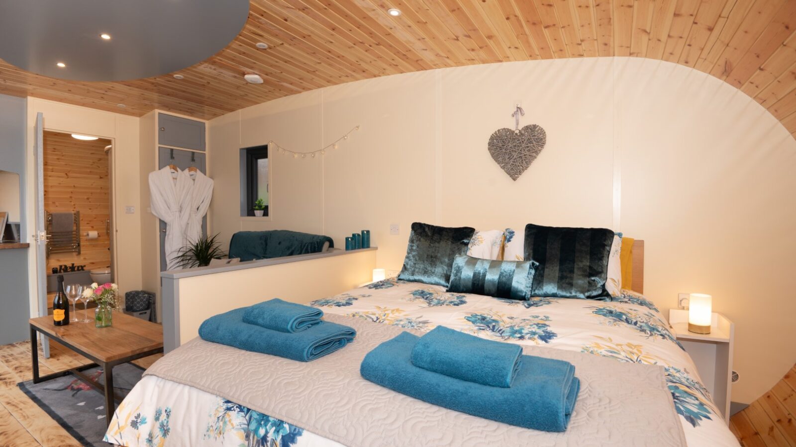 A cozy bedroom at Beacon View Pods features a wooden ceiling, a double bed with teal accents, two folded towels, and a small table set for drinks.