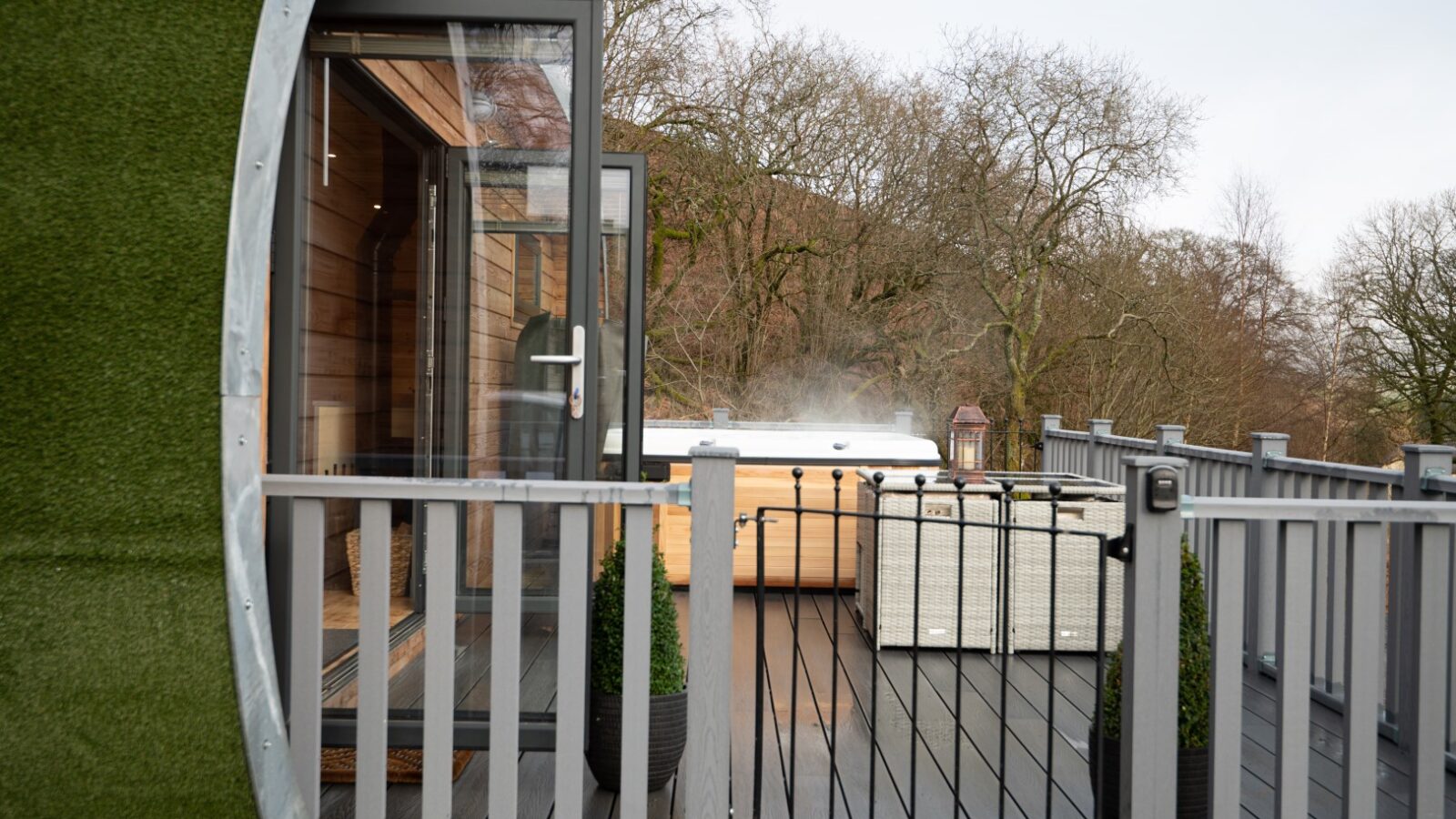 The cosy outdoor deck of the Beacon View Pods features a glass door entrance, two planters, and a hot tub surrounded by a wooded area. Perfect for glamping, the steaming hot tub offers a peaceful retreat with bare trees in the background.