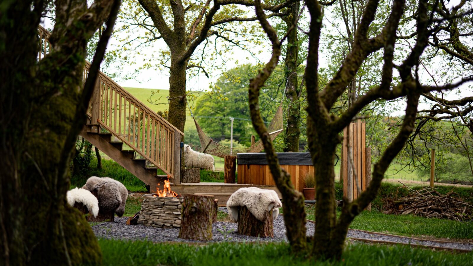 A cozy outdoor setting featuring a wooden hot tub and a stone fire pit surrounded by log seats with fur covers. Enclosed by tall, leafy trees, this Welsh-inspired retreat includes a wooden staircase leading to treehouses on an elevated platform in the background.