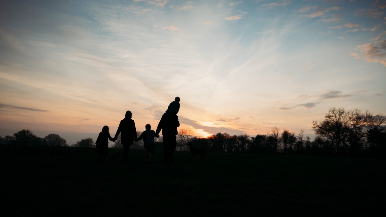 Silhouette of a family holding hands while strolling past Pennard Farm at sunset. The sky is a mix of orange, pink, and blue hues, with scattered clouds. Trees line the horizon, adding to the tranquil scene by the cottages.