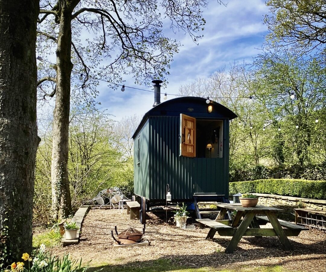 A small, green shepherd's hut with a chimney is set in a wooded area. Perfect for a serene getaway, this Eyam accommodation offers cozy charm. Nearby, there's a picnic table and string lights to complete the enchanting setting of your ideal hut rental experience.