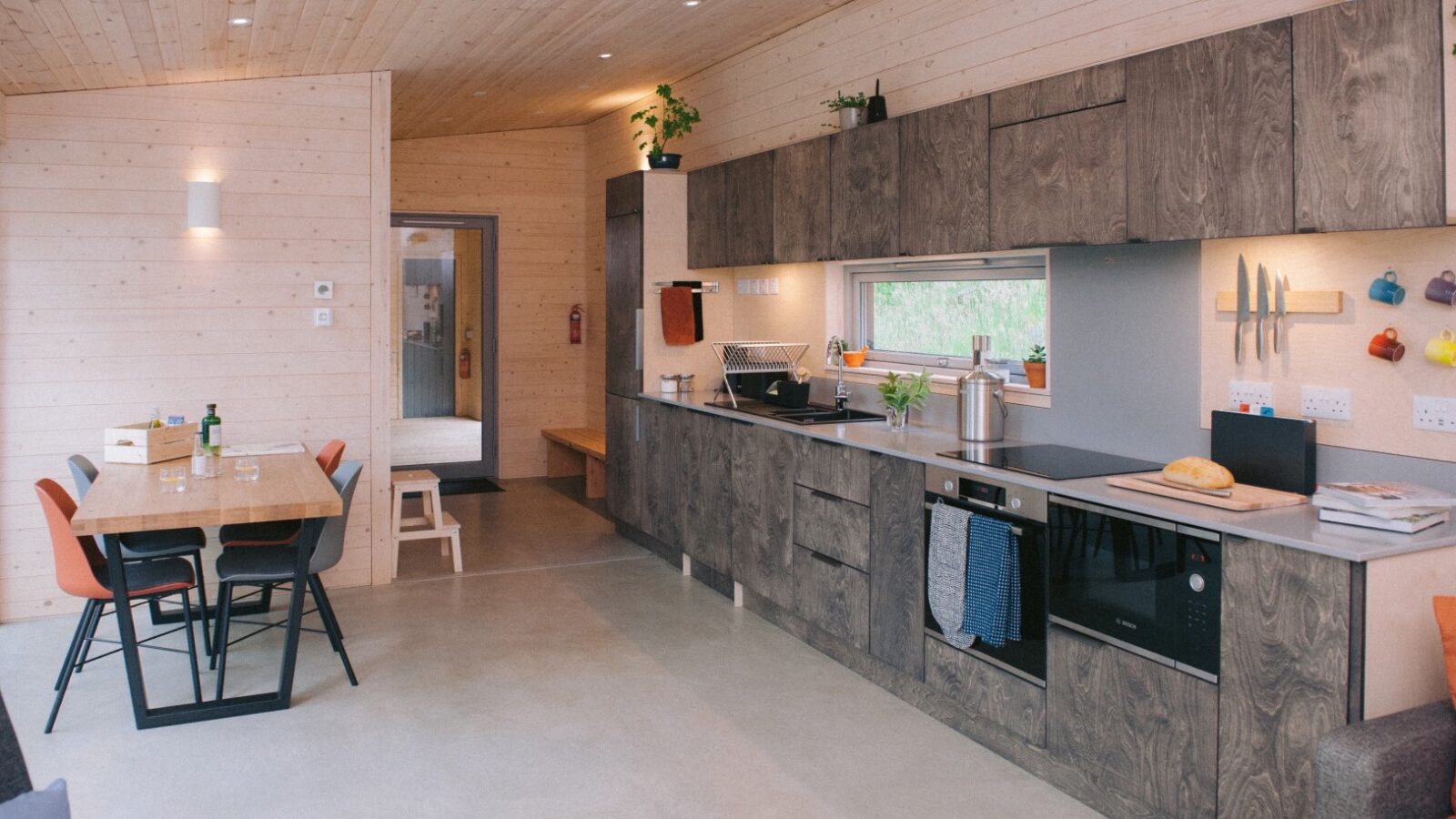 A modern kitchen in Ecotone Cabins boasts wooden wall panels and cabinetry. The space features a dining table with colorful chairs, a countertop with a sink, a stove, and a window. Knives and utensils are mounted on the wall, while potted plants provide refreshing greenery.