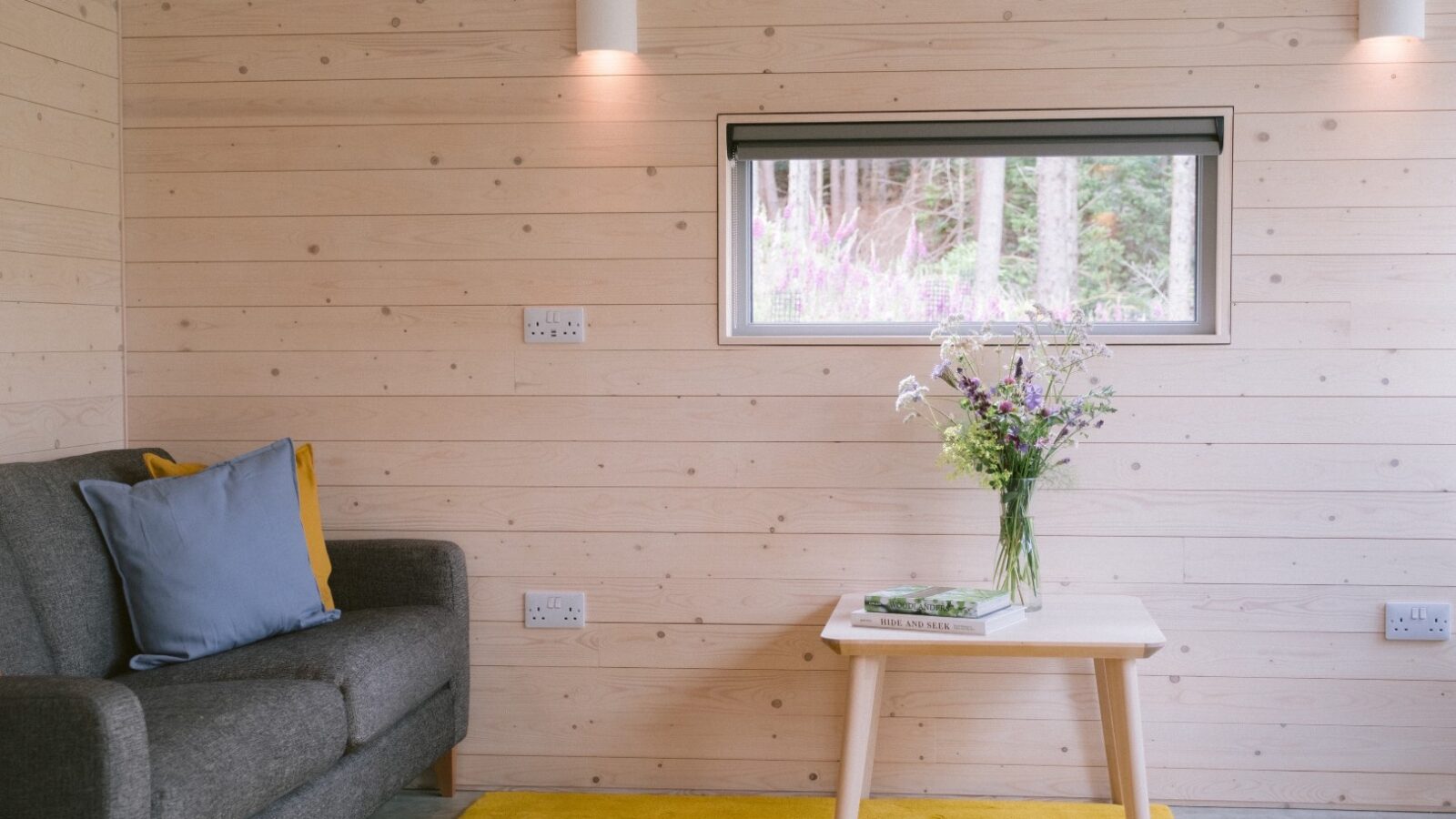 A modern living room in the style of Ecotone Cabins features light wood panel walls and a small window. A gray sofa with blue and yellow cushions, a wooden table with a vase of wildflowers, and a yellow rug adorn the floor. Wall lights illuminate this cozy, cabin-inspired space.