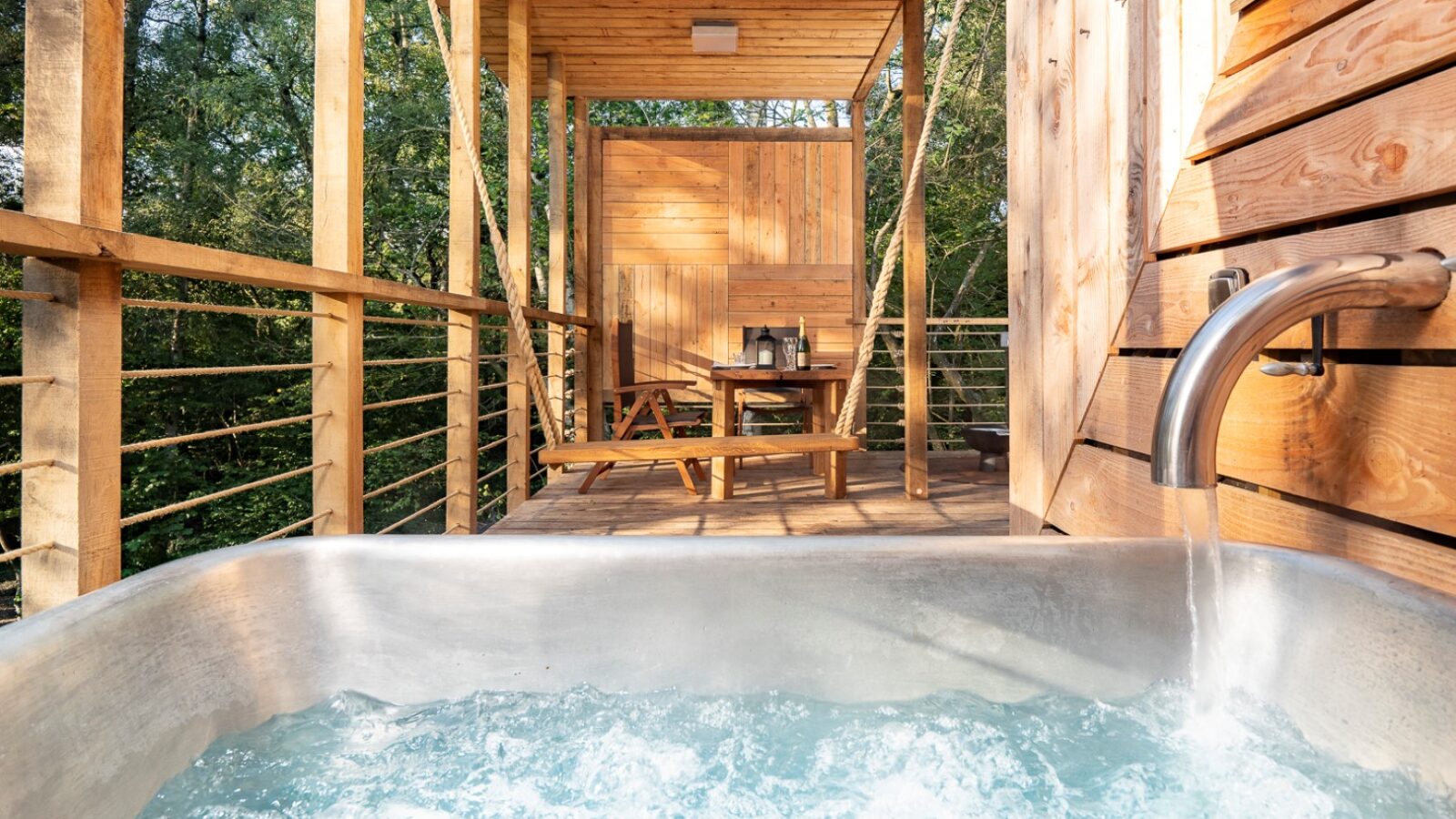 A wooden balcony set amidst trees features a hot tub with bubbling water in the foreground. In the background, there is a wooden picnic table and chairs, creating a serene and rustic outdoor setting reminiscent of the charming Pinwheel Treehouse.