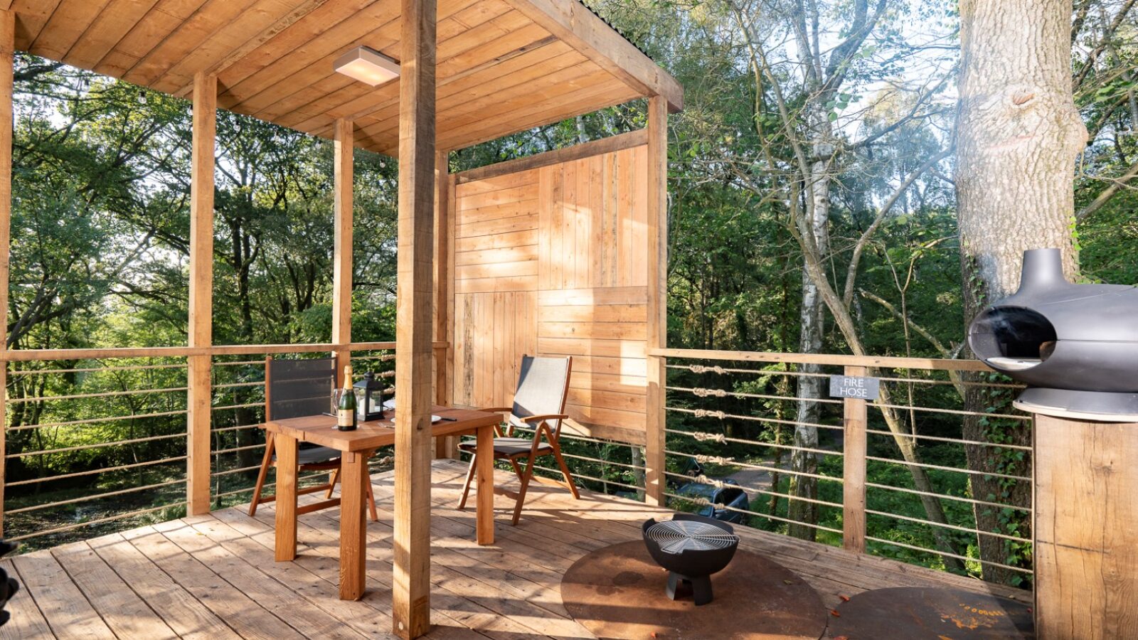 A cozy wooden deck nestled among tall trees, resembling a charming treehouse, features a small table with two chairs, a reclining chair, a modern cylindrical fireplace, and a partial wooden canopy providing shade. Soft sunlight filters through the surrounding greenery, creating a serene atmosphere.