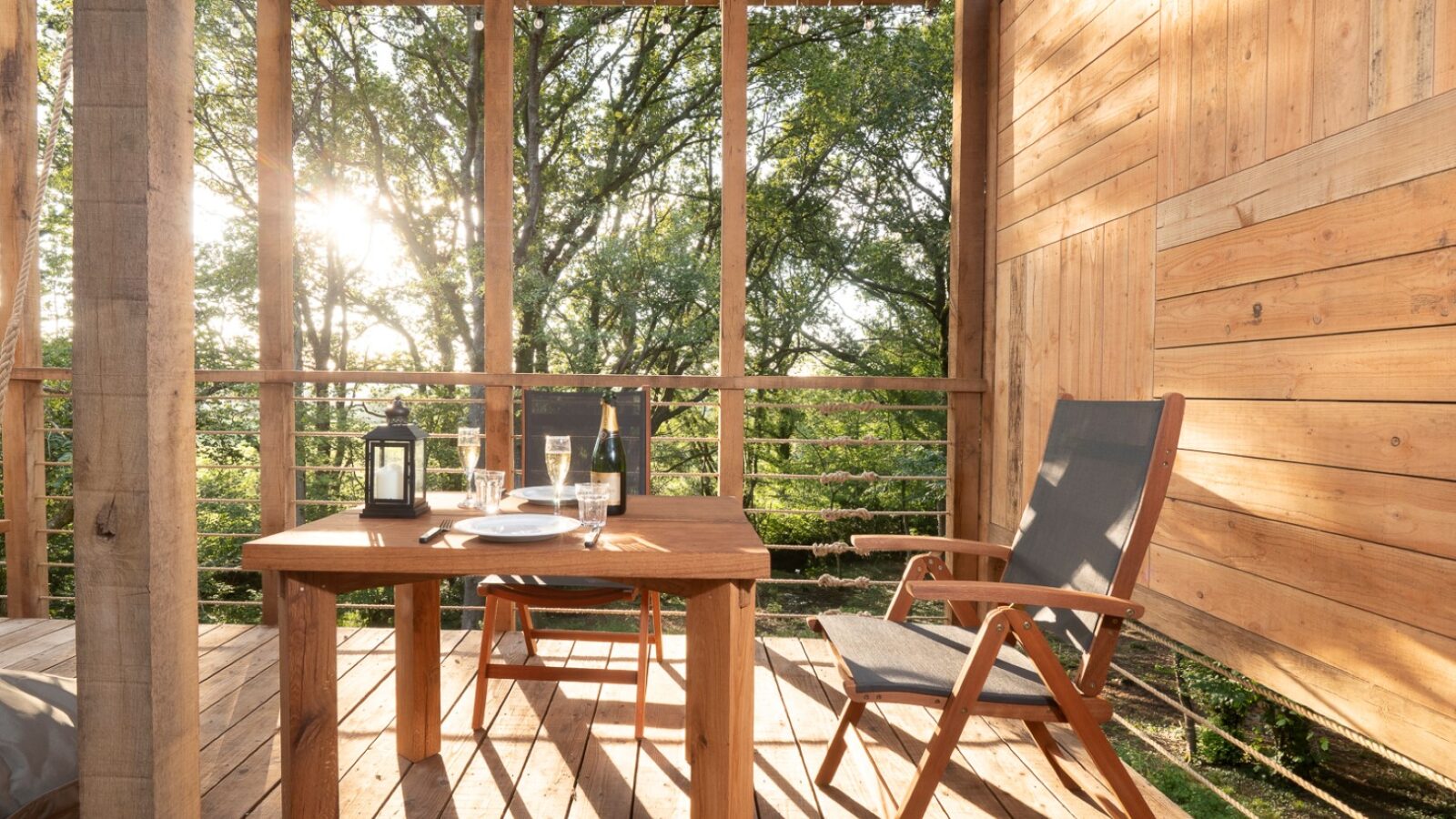 A wooden outdoor balcony with two chairs and a table set for two, featuring a lantern, drink glasses, and plates. The treehouse-style balcony overlooks lush green trees, with sunlight streaming through the foliage, creating a serene and cozy atmosphere.