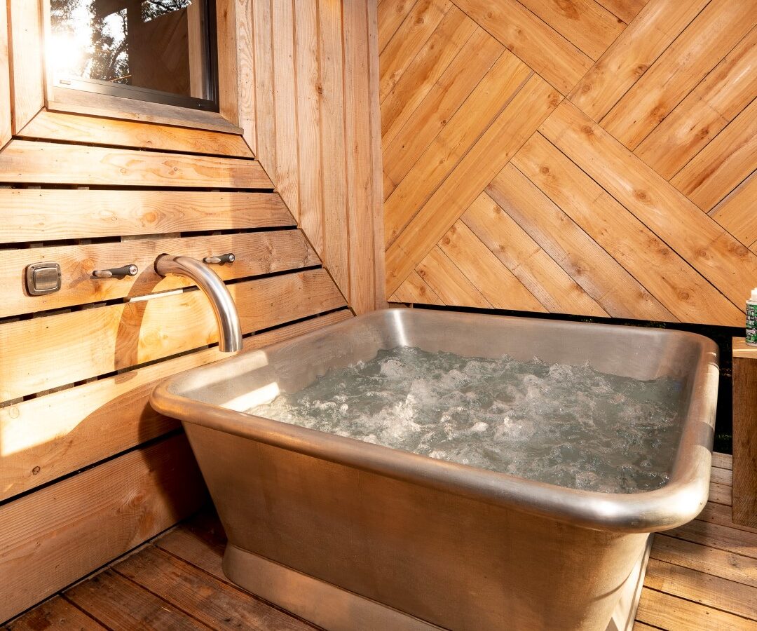 A modern outdoor wooden deck, reminiscent of a chic treehouse, features a luxurious rectangular stainless steel soaking tub filled with water. Set against a stylish wooden wall with diagonal planks, the serene space is complemented by a window offering views of the surrounding nature.