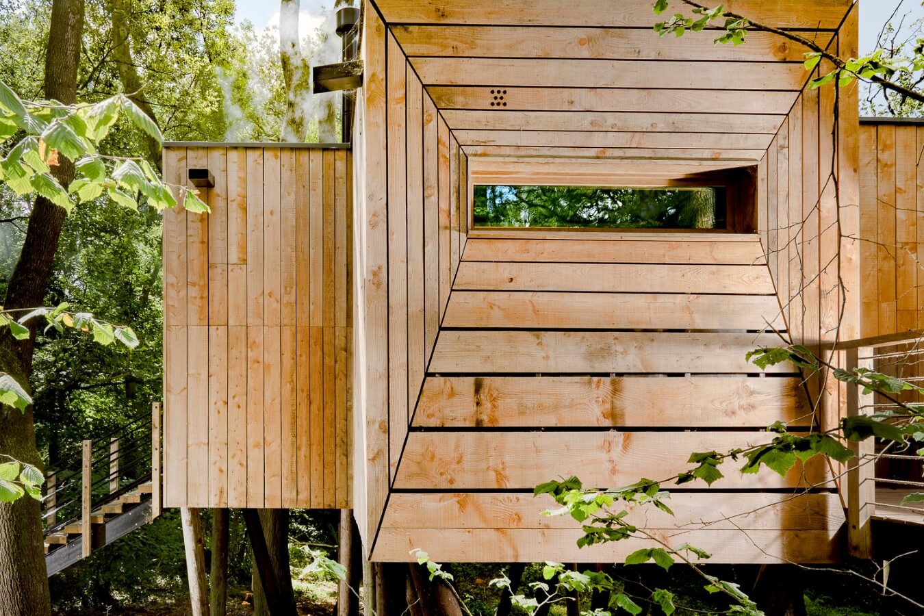 A modern wooden treehouse built among trees features a rectangular window and siding. Green foliage surrounds the treehouse, with a pinwheel spinning in the gentle breeze by the walkway on the left. Sunlight filters through the trees, adding a warm glow to the scene.