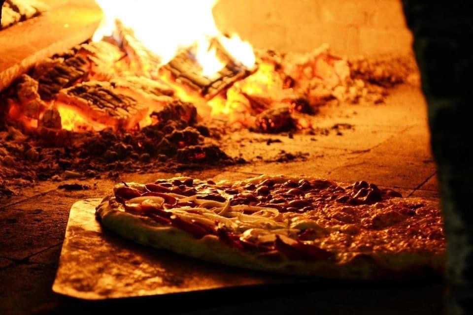 A pizza on a peel is being baked in a wood-fired oven, with flames flickering in the background—perfect for cozy woodland escapes.